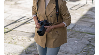 photographer wearing light brown jacket and jeans carrying Sony a7R camera with FE 24mm G Master Lens