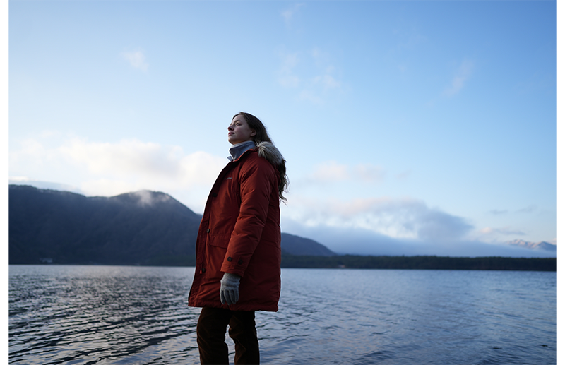 Sony FE 24mm F1.4 GM Lens sample image portrait of woman wearing orange winter coat standing infront of large lake with mountains in the background